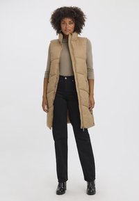 Gilet rembourré sans manches couleur khaki avec un col haut, fermeture éclair et poches latérales, porté par-dessus un haut à manches longues beige et un pantalon noir.