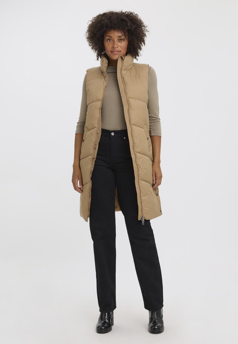 Gilet rembourré sans manches couleur khaki avec un col haut, fermeture éclair et poches latérales, porté par-dessus un haut à manches longues beige et un pantalon noir.