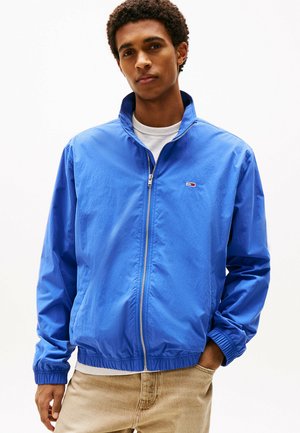 Blauwe lichtgewicht zipjas met een staande kraag, elastische manchetten en zoom. Bevat een klein logo op de borst. Draagt over een wit shirt.