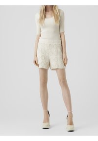 Bílý pletený top s krátkými rukávy, který je doplněný krémovými krajkovými šortkami s diamantovým vzorem a třásněmi, k tomu krémové platformové podpatky.
