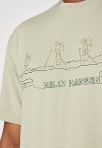 Světle zelené tričko vyrobené z měkké látky s liniovým výkresem horolezců a textem "HELLY HANSEN" ve tmavší zelené barvě na spodní části.