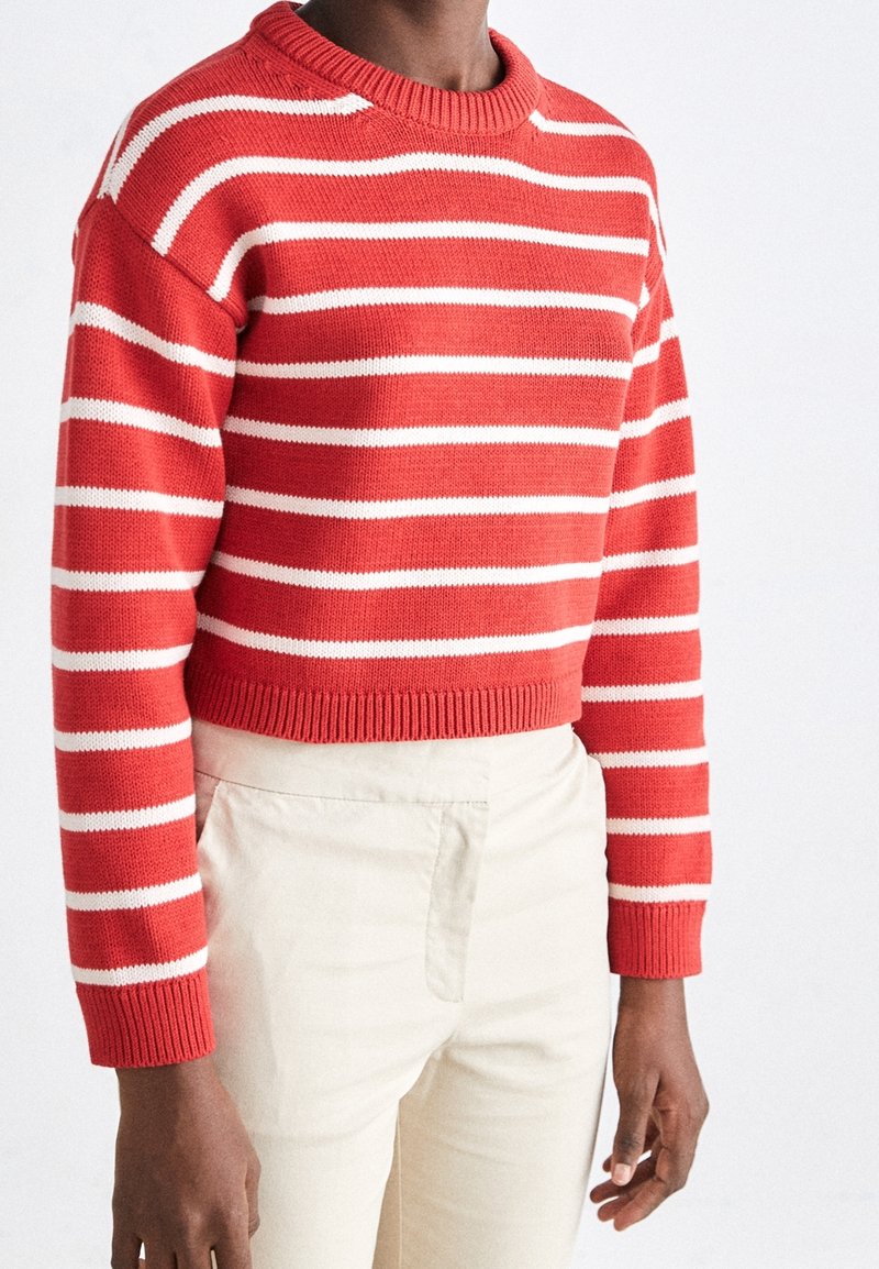 Pull en tricot rayé rouge et blanc avec un col rond et des épaules tombantes ; poignets et ourlet côtelés. Associé à un pantalon beige clair.