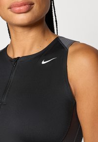 Haut de sport noir Nike avec fermeture éclair et accents gris texturés sur les épaules. Matière lisse et extensible avec une coupe ajustée.