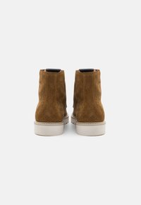 Botines de ante marrón con textura suave, suelas de goma blanca y diseño minimalista. Disponen de una pestaña en la parte trasera para facilitar el uso.
