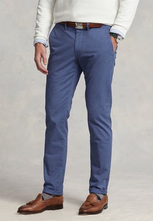 Άνδρας που φορά μπλε chinos, καφέ δερμάτινη ζώνη, καφέ loafers με φούντες, γκρι κάλτσες με σχέδια, λευκό πουλόβερ και ρολόι χειρός, στέκεται με το ένα χέρι στην τσέπη.