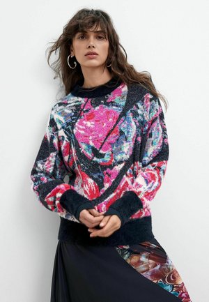 Kvinde med langt bølget hår iført øreringe i hoop, en farverig blomstret sweater og et sort skørt med et blomstret panel.