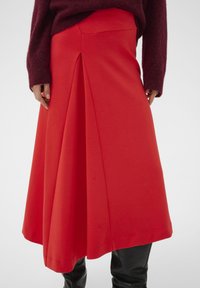 Roter plissierter Rock aus glattem Stoff, mit breitem Saum und dezenten Nahtdetails, getragen zu einem dunklen Pullover.