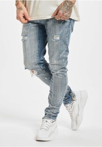 Lyseblå revne jeans med slitte detaljer, tettsittende design og frynsete kanter. Kombinert med hvite joggesko med teksturerte aksenter.