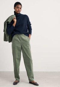 Mornarska polovična majica s turtleneck ovratnikom, rebrast tekstur, v kombinaciji s svetlo zelenimi karirastimi hlačami. Model stoji z zelenim jopičem obešenim čez eno ramo.