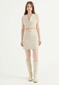 Top corto beige chiaro con bottoni, abbinato a una minigonna coordinata, entrambi con motivo a spina di pesce. Stivali beige fino al ginocchio.