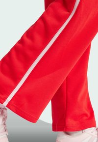 Crvene trenirke s bijelim bočnim prugama, mekog materijala i opuštenog kroja, u kombinaciji s svijetloružičastim sportskim cipelama.