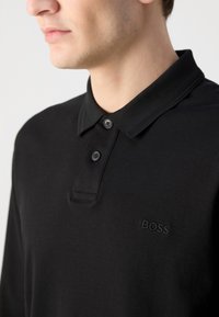 Juodas polo marškinėlis, pagamintas iš minkštos medvilnės, turintis klasikinį apykaklę, du juodus mygtukus ir išsiuvinėtą "BOSS" logotipą krūtinėje.