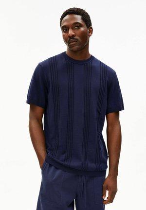 Erwachsener Mann mit kurzen lockigen Haaren und Schnurrbart, der ein marineblaues, kurzärmeliges Strickhemd und passende marineblaue Hosen vor einem einfarbigen Hintergrund trägt.