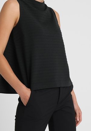 Sort ærmeløs top med diskrete horisontale striber parret med sorte bukser, den ene hånd i lommen, mod en ensfarvet lys baggrund.