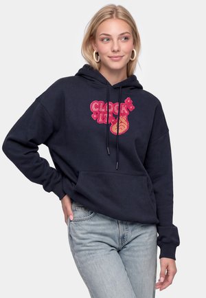 Ung kvinde med blondt hår iført øreringe i ringe, en navy hoodie med pink grafisk tekst og lyseblå jeans, poserer med den ene hånd på hoften.