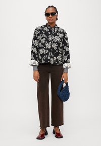 Giacca nera con motivo floreale e fiori bianchi, polsini a coste grigi, pantaloni dritti marroni, scarpe con tacco a blocco rosse e borsa intrecciata blu.