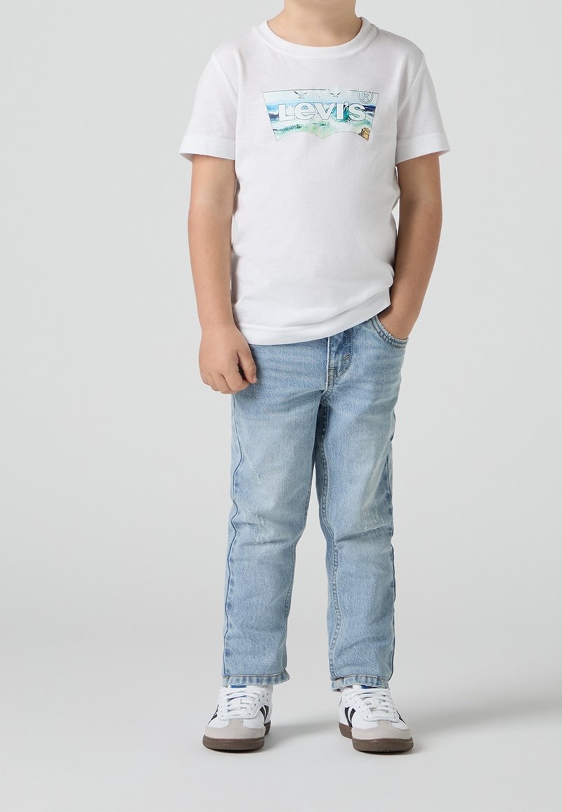 Enfant portant un t-shirt blanc Levi's, un jean bleu clair et des baskets blanches, debout avec une main dans la poche sur fond uni.