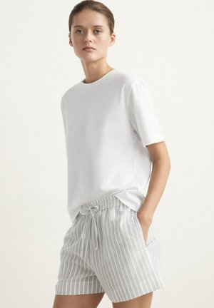 Ung kvinde står iført en simpel hvid kortærmet T-shirt og lysegrå shorts med striber og snøre, kigger fremad med hånden i lommen.