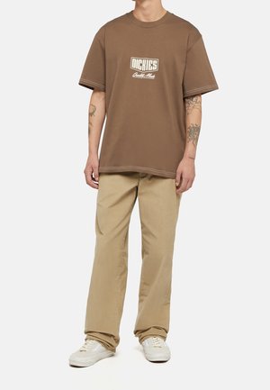 Camiseta marrón de manga corta con cuello redondo y gráfico de logo, combinada con chinos beige claro y zapatillas blancas. Diseño sencillo, ajuste relajado.