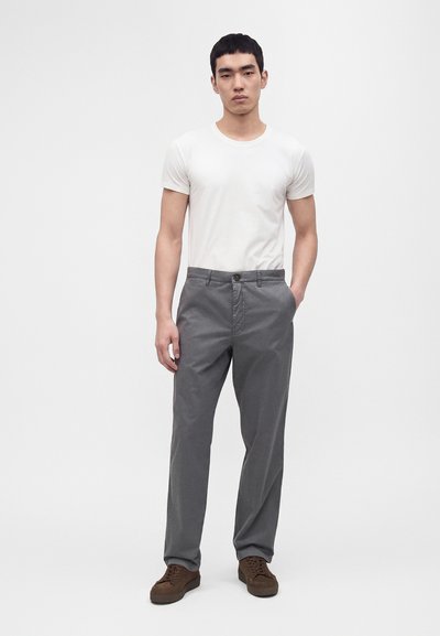 Homme portant une chemise blanche à manches courtes, un pantalon gris et des baskets marron, debout avec une main dans la poche devant un fond uni.