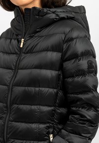 Zwarte pufferjack met een ritsluiting, capuchon en horizontaal gestikt ontwerp. Voorzien van gouden hardware en een logo op de linker mouw.