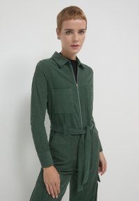Combinaison en velours côtelé vert avec une fermeture éclair sur le devant, un col, des manches longues, deux poches avant et une ceinture nouée à la taille. Tissu texturé à côtes.