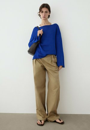 Vrouw staat tegen een witte muur, draagt een blauwe trui, losse beige broek, zwarte schoudertas en zwarte sandalen.