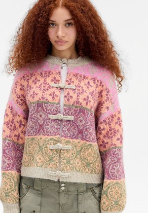 Giovane donna con capelli rossi ricci che indossa un cardigan lavorato a maglia multicolore con motivi floreali e chiusure a fettucce, abbinato a pantaloni verde oliva.