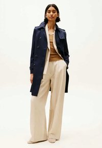 Ναυτικό trench coat από λείο ύφασμα, σε συνδυασμό με μπεζ παντελόνι σε φαρδιά γραμμή. Το μοντέλο φοράει μια ανοιχτόχρωμη μπλούζα και παπούτσια με σχέδιο. Απλή, καθαρή σιλουέτα.