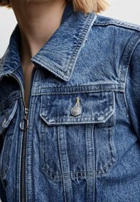 Denimjakke med glidelås, brystlomme med klaff og metallknapp, båret av en person med kort, blond hår synlig ved kragen.