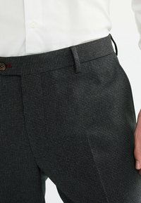 Pantalons gris texturé avec un petit motif à carreaux, dotés d'une fermeture à bouton et de passants de ceinture, positionnés sur un fond clair.