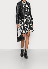 Osoba v černých květovaných šatech s předním rozparkem, černé kožené bundě, černých kotníkových botách a držící černou koženou kabelku.