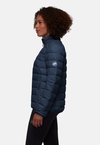Marineblauwe pufferjack met rits, met een hoge kraag, gestikte uitstraling en een logo op de linkermouw. Gekleed met zwarte leggings.