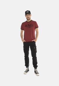 T-shirt rouge avec des accents noirs, texte du logo sur le devant, associé à un pantalon cargo noir et des baskets noires avec des lacets blancs.