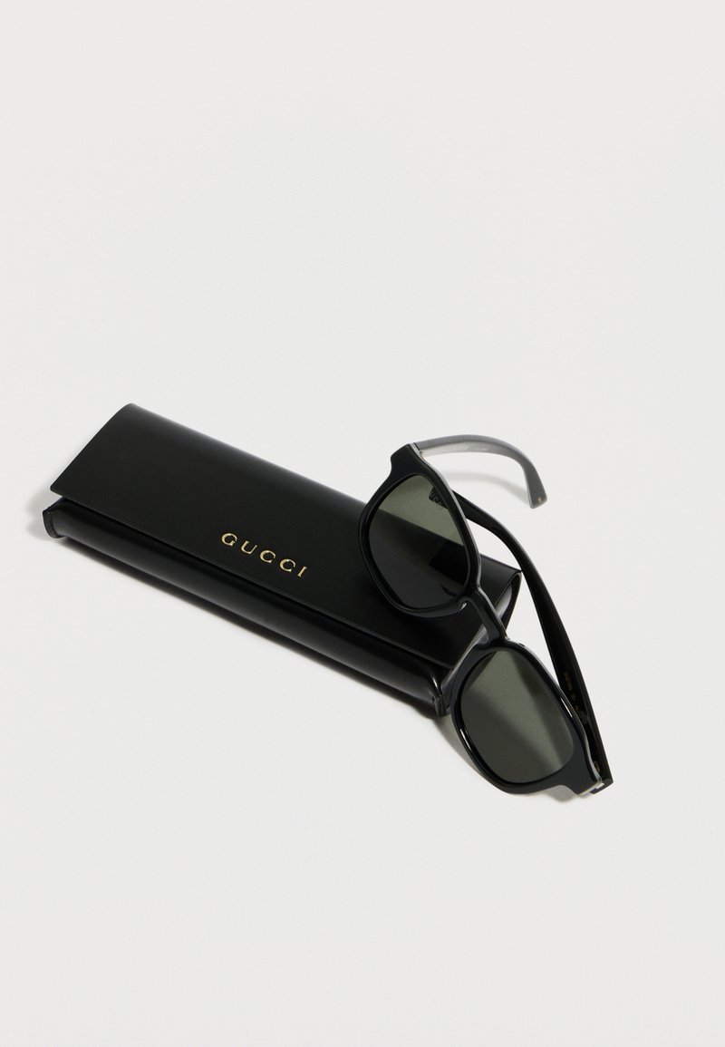 Lunettes de soleil Gucci rectangulaires noires avec des verres foncés reposant sur un étui Gucci noir assorti sur un fond blanc.