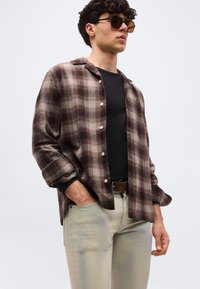 Άνδρας που φορά καφέ καρό πουκάμισο με κουμπιά πάνω από μα ύρο μπλουζάκι, ανοιχτό μπεζ παντελόνι και σκούρα γυαλιά ηλίου, με το ένα χέρι στην τσέπη.