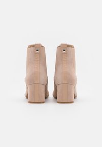 Botas de tobillo de ante beige con puntera redondeada, tacón cuadrado y paneles laterales elásticos. Cuenta con un lazo en la parte trasera y detalles de costura vertical.