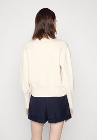 Cremfarbener Pullover mit Puffärmeln und geripptem Saum, kombiniert mit dunklen Marineblau-Shorts. Weiche Stricktextur. Rückansicht gezeigt.