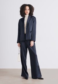 Blazer navy e pantaloni a gamba larga in tessuto liscio, abbinati a un dolcevita crema. Accessoriati con una piccola borsa bianca e tacchi neri.