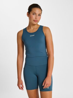 Ung kvinde iført blå ærmeløs sportstop og matchende højtsiddende shorts, står mod en ensfarvet lys baggrund.