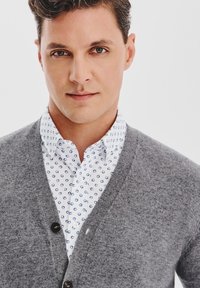 Mężczyzna z krótkimi brązowymi włosami w białej koszuli z niebieskim wzorem i szarym kardiganie, patrzy bezpośrednio w kamerę na białym tle.