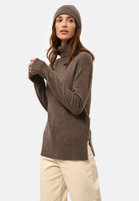 Brun ullturtlenecktröja med ribbade muddarna och sidofickor, parat med en matchande mössa. Modellen poserar mot en enkel bakgrund.