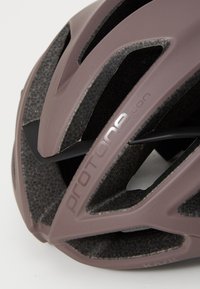 Aerodynamiczny kask rowerowy w stonowanym mauve z teksturowanymi czarnymi akcentami. Posiada wyraźne otwory wentylacyjne oraz detal z logo "protone".