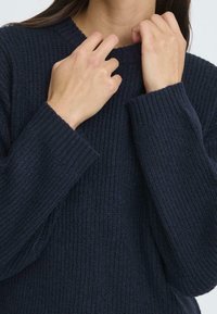 Sötétkék, kötött pulóver bordás textúrával, széles mandzsettákkal és magas gallérral. Az anyag puhanak és kényelmesnek tűnik, alkalmas hétköznapi viseletre.