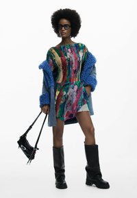 Többszínű, absztrakt mintás ruha laza szabással, réteges farmerkabát bolyhos kék mandzsettákkal, térdig érő fekete csizmák, és kis fekete táska.