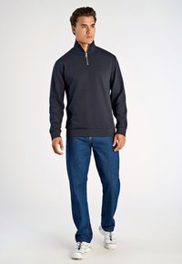 Sudadera de cuello con cremallera en color navy con puños y dobladillo acanalados, combinada con jeans azules y zapatillas deportivas blancas, de pie frente a un fondo liso.