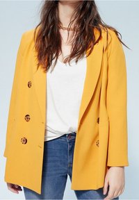 Blazer cruzado de tela amarilla brillante con grandes botones marrones, combinado con una camisa blanca y jeans azules. Textura suave, corte ajustado.