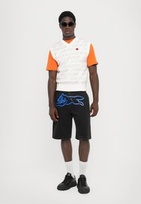 Top de punto gráfico blanco y naranja con el texto "HELADO", combinado con pantalones cortos de mezclilla negros que presentan un contorno de perro azul. Zapatillas negras y calcetines blancos.