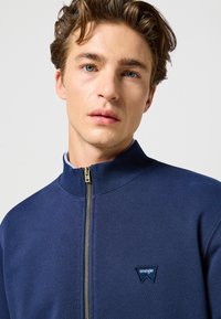 Marinblå zip-up sweatshirt i mjukt tyg, med hög krage, guldfärgad dragkedja och en liten blå broderad Wrangler-logga.