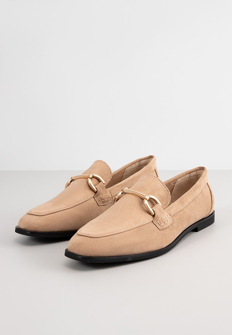 Beige Wildleder Slipper mit einer niedrigen schwarzen Sohle, die ein goldfarbenes Akzentband an der Vorderseite und eine glatte, strukturierte Oberfläche aufweisen.